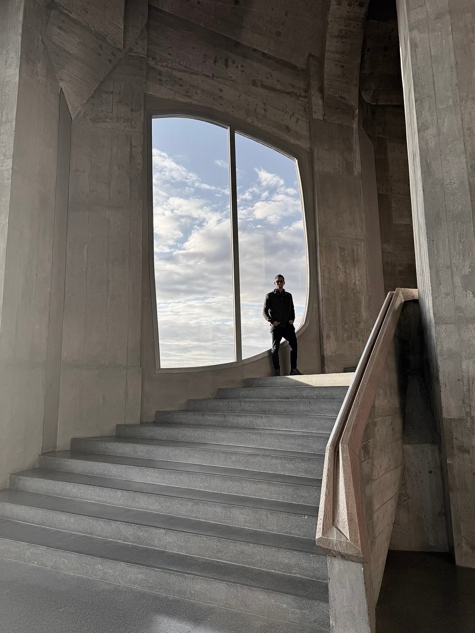Sky Mathis standing at the head of a concrete stair, framed by a tall curved window opening onto cloud and sky.